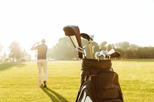 Bolsa de golf en primer plano con muchos palos y un hombre en la distancia golpeando.
